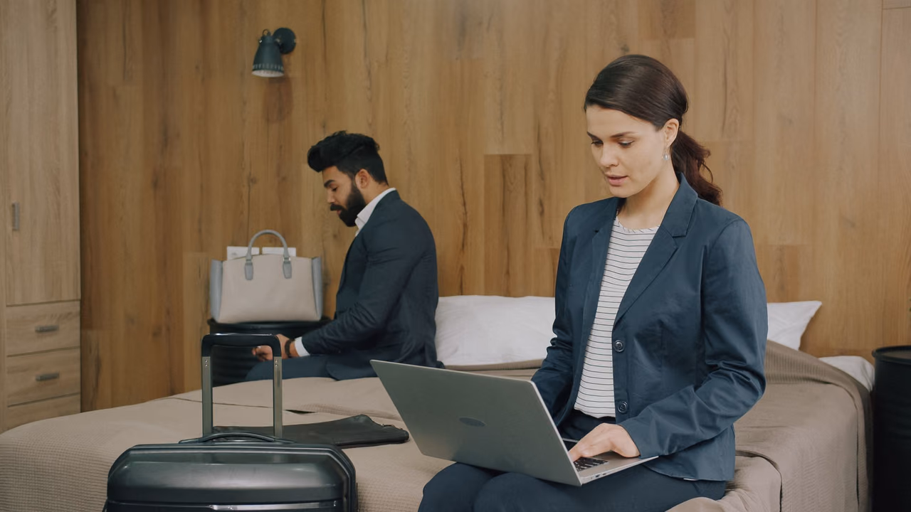 Two business travelers in blazers on a hotel bed, one typing on a laptop, with a rolling suitcase in the foreground
