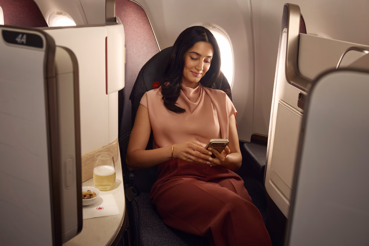A passenger looks at a phone in a window seat in Air Canada business class, with a drink and snack on the side table.