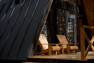 Two wooden Adirondack chairs on the sunlit porch of a dark A-frame cabin