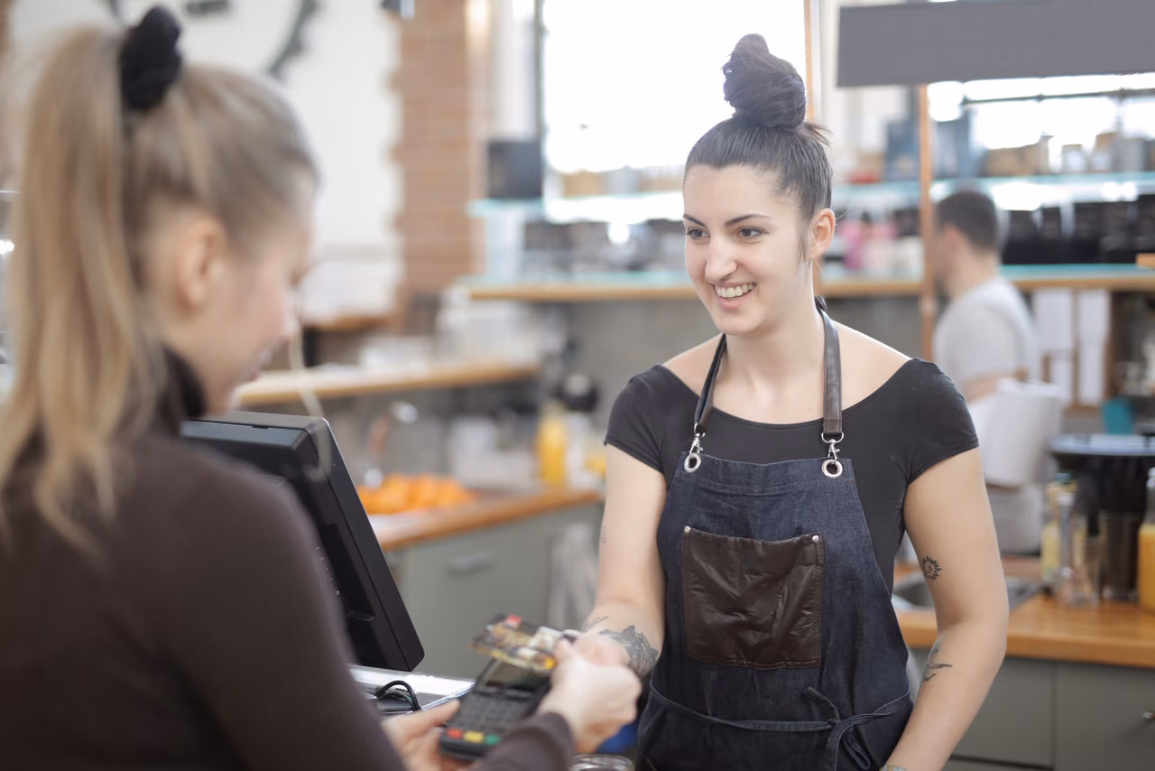 A customer taps a card on a payment terminal while a smiling barista in a denim apron stands behind a cafe counter