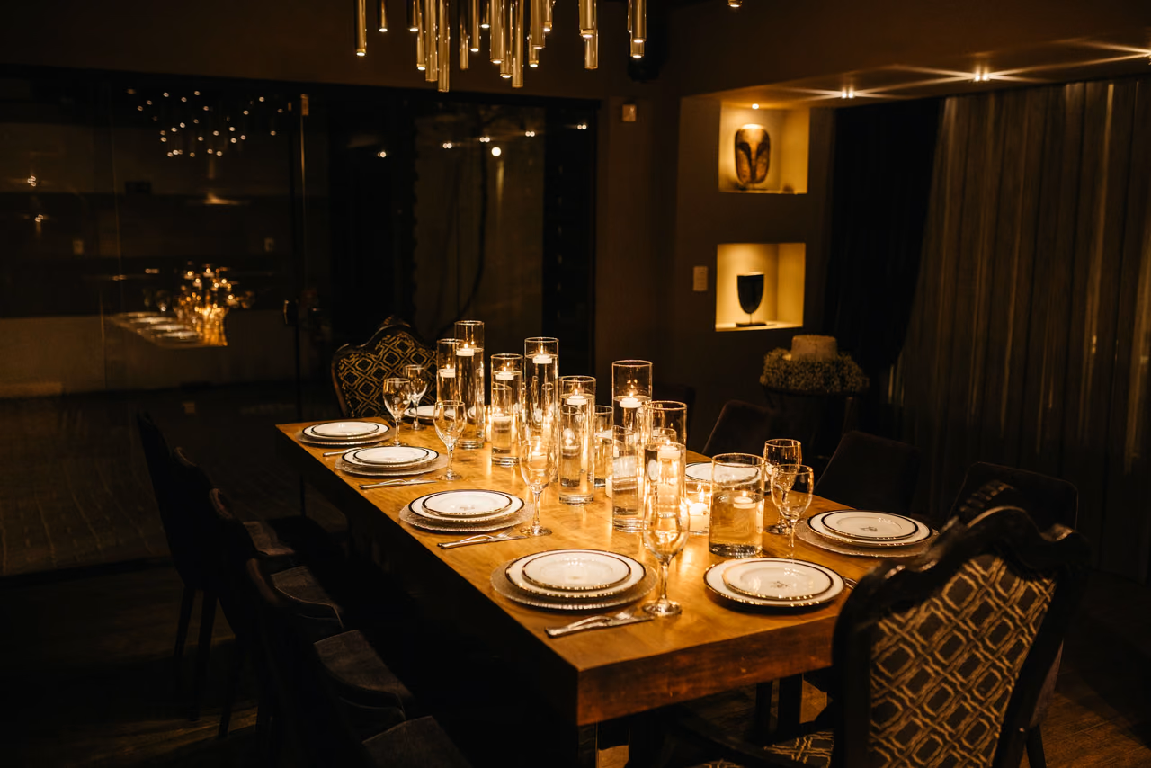 A long wooden dining table set with white plates, wine glasses and rows of lit candles under a hanging chandelier in a dim private dining room.