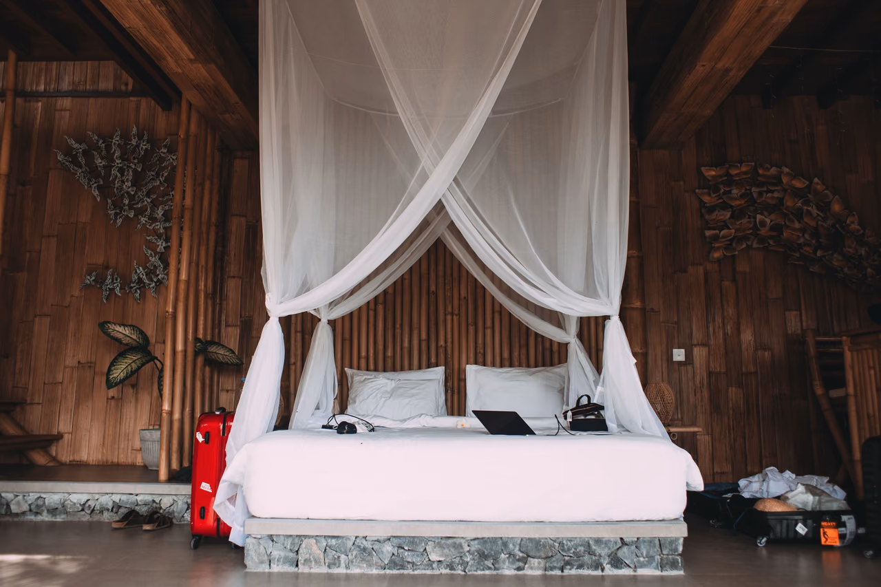 A white canopy bed with a laptop on it in a wood-paneled vacation room, with a red suitcase on one side and an open bag on the other.