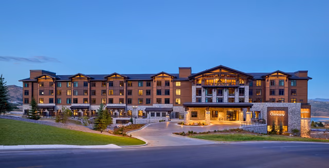 The five-story Canopy by Hilton Deer Valley at dusk, with lit windows, a stone and timber entrance and a Canopy sign, against hills and a clear blue sky.