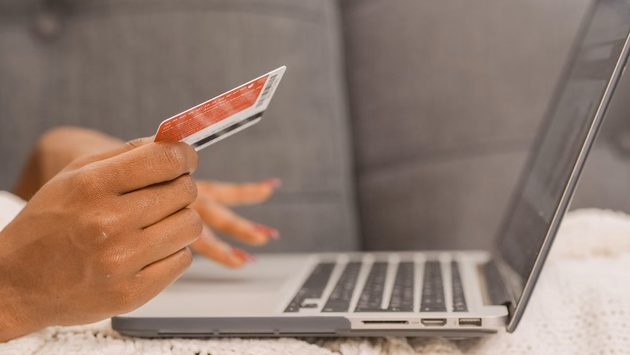 A hand holds a red credit card beside an open laptop on a white knit blanket