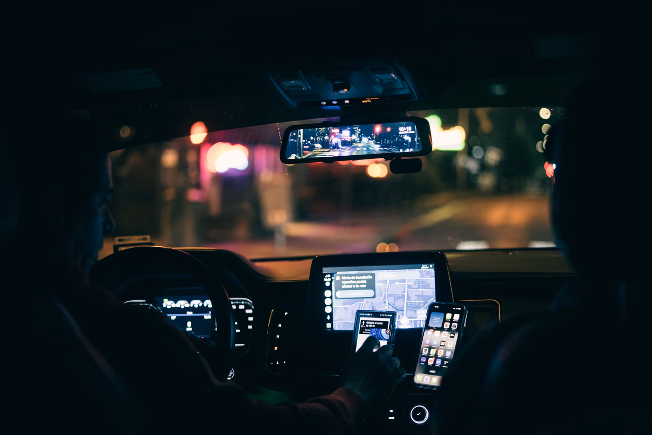 View from the back seat of a car at night, with a driver and two phones mounted beside a navigation screen as city lights blur past the windshield