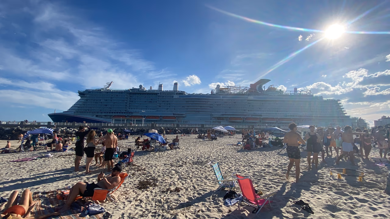 The cruise ship Carnival Celebration passing a crowded Miami beach under a bright sun, beachgoers in chairs in the foreground