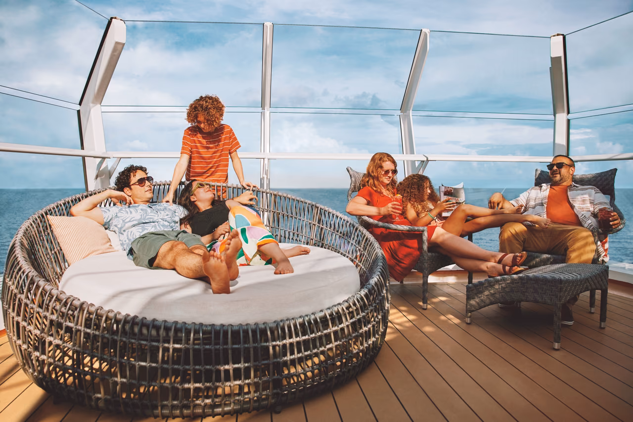 A family on a Carnival ship's open deck, two in a round wicker daybed, a child behind it and three in deck chairs by the sea
