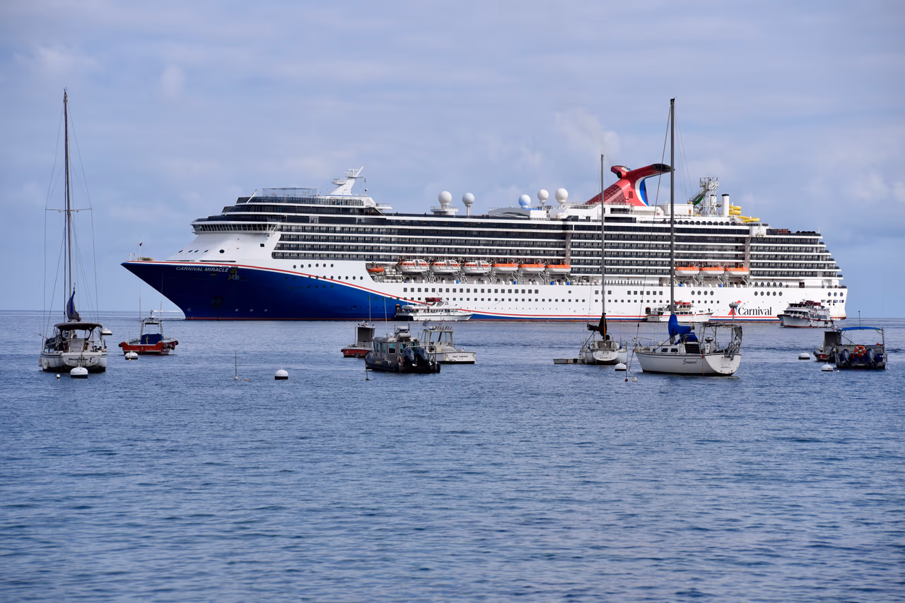 A Carnival cruise ship at anchor offshore under broken cloud, with sailing boats and small craft moored between the ship and the camera