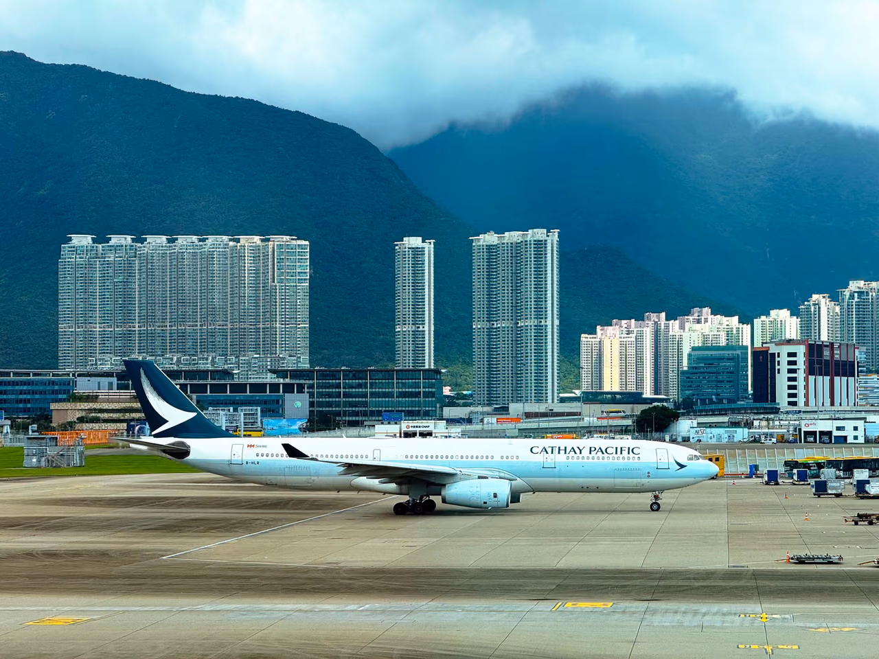 A Cathay Pacific Airbus A330 taxiing at Hong Kong International Airport, with apartment towers and cloud-topped green mountains behind