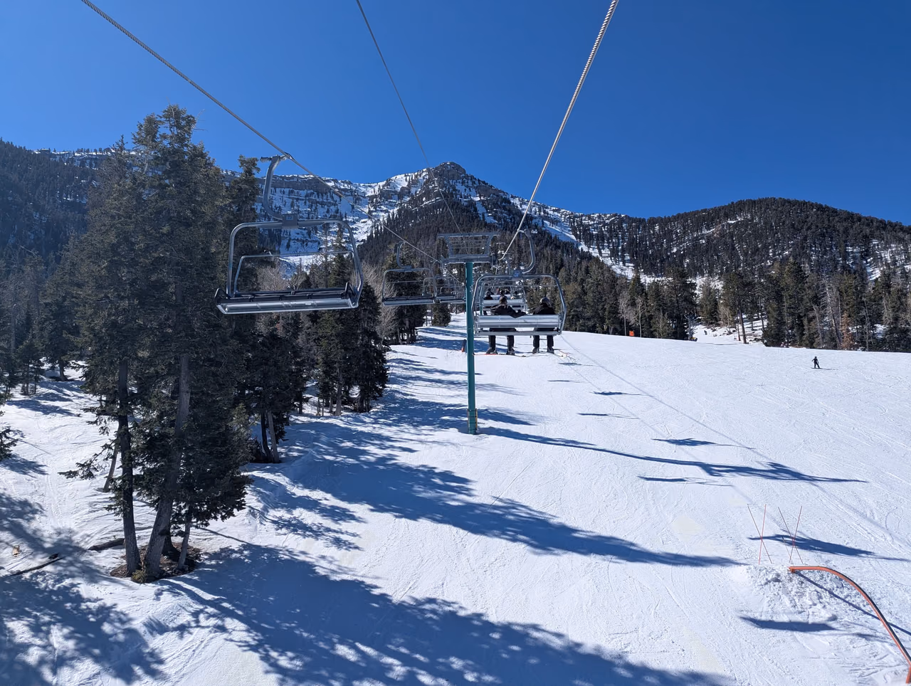 A chairlift with a few riders climbs over a groomed snowy slope lined with evergreen trees toward a mountain ridge under a clear blue sky.