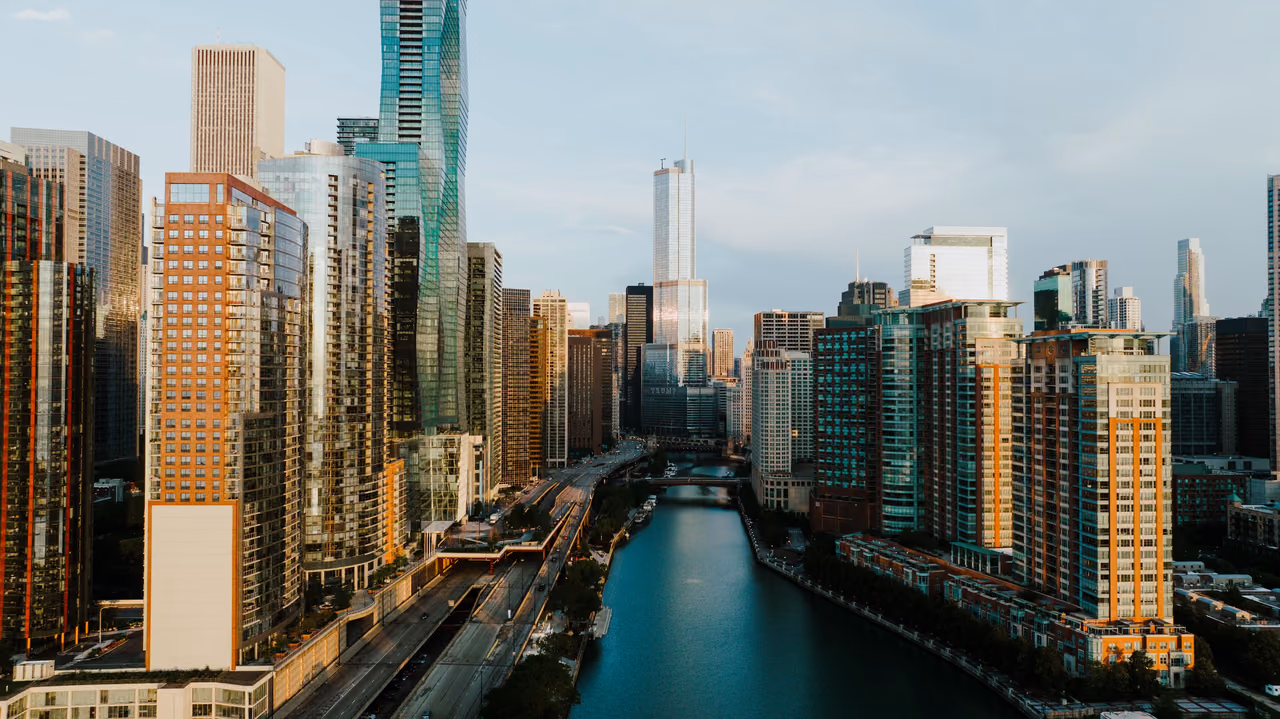 An aerial view of the Chicago River running between high-rise towers in downtown Chicago, with the skyline lit by low evening sun