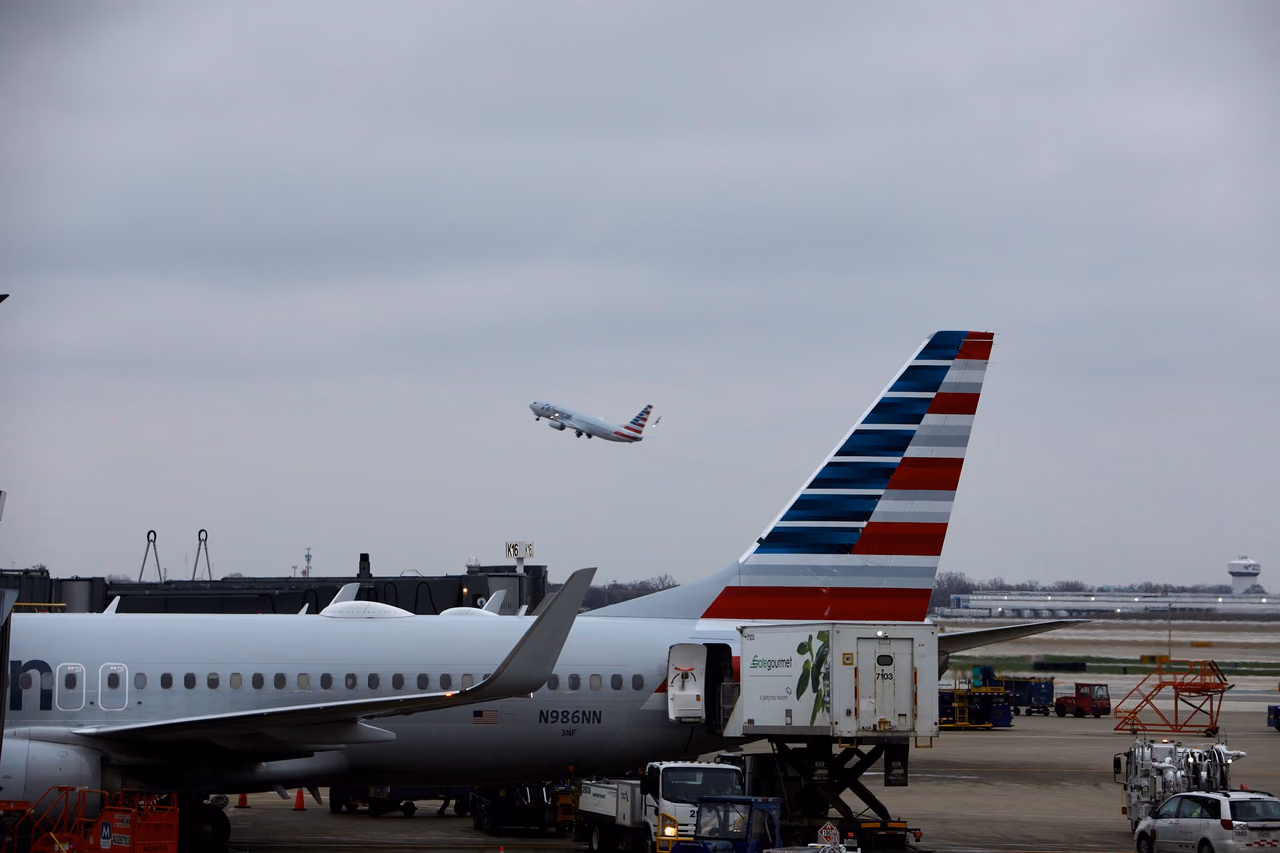An American Airlines jet takes off behind a parked American Airlines aircraft being loaded by a catering truck at the gate.