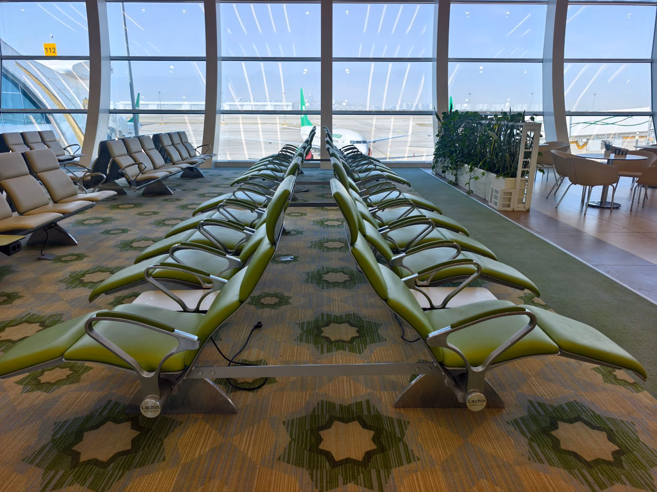 Rows of green lounge chairs in an airport terminal with floor-to-ceiling windows and an aircraft parked outside