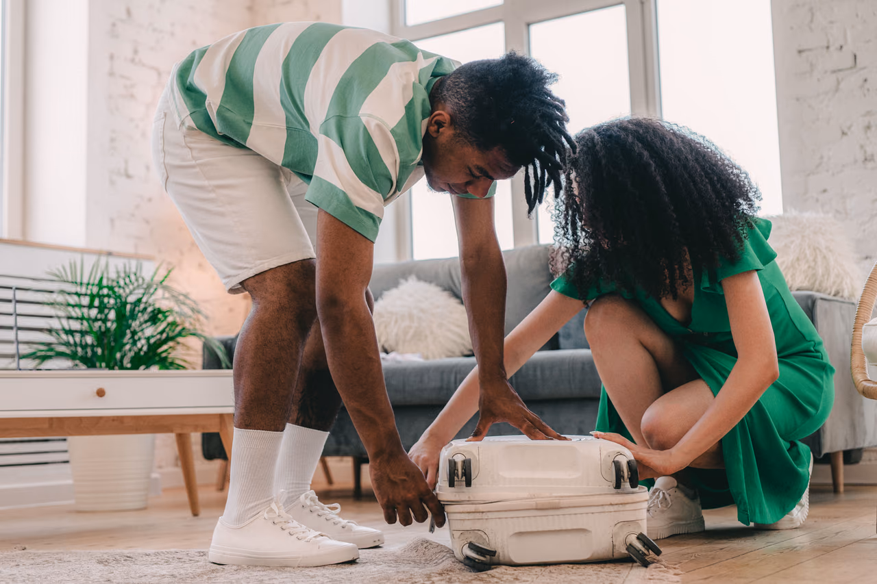 A couple in a bright living room pressing down together to close a white hard-shell suitcase