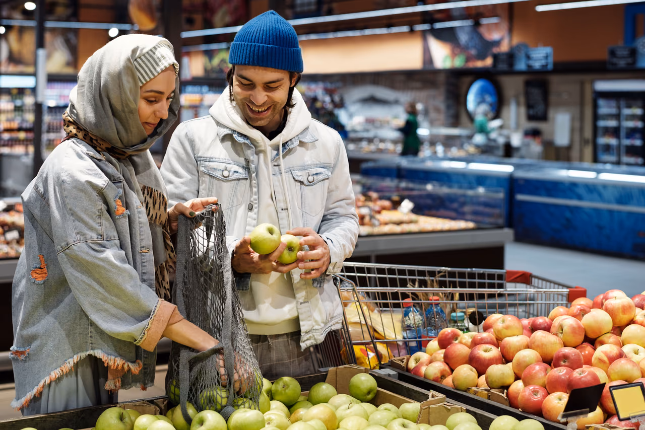 A couple picks out apples together in a supermarket produce section with a shopping cart beside them.