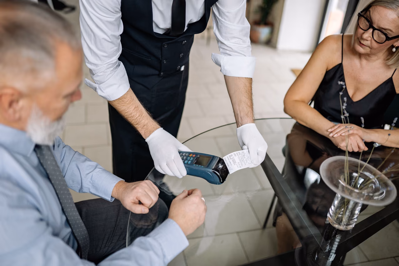 A waiter in white gloves holds a card terminal and receipt over a glass table while a couple settles the bill at a restaurant