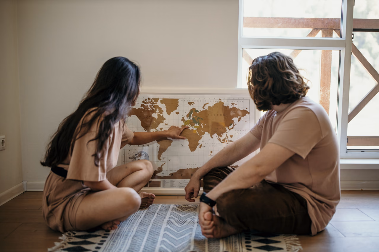 A couple sits on a wooden floor pointing at a world map propped against the wall, planning where to travel next.