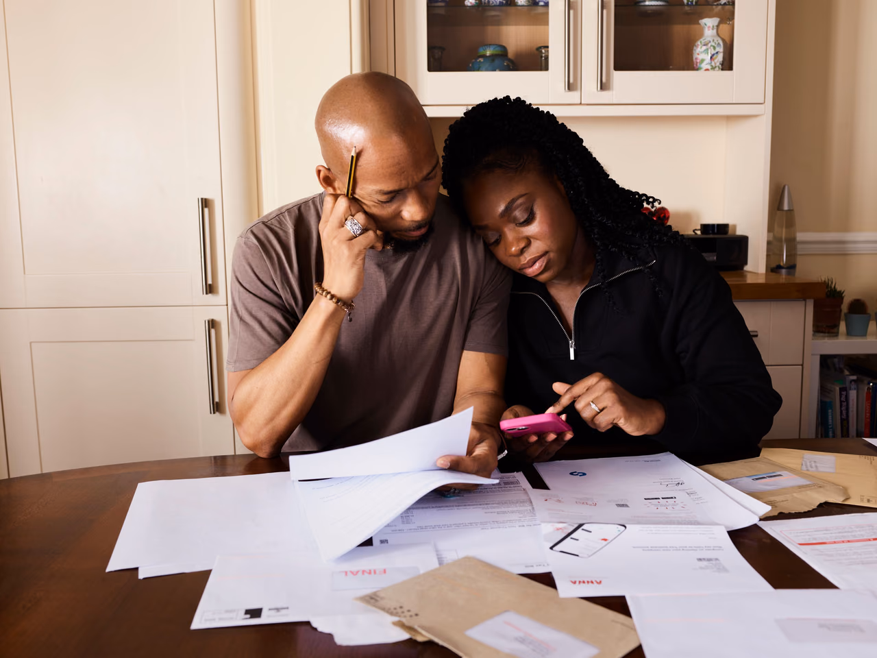 A couple at a dining table covered in bills and final-notice letters, the man holding papers and the woman using a calculator.