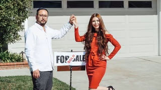 A couple stands hand in hand in front of a house beside a for-sale sign marked sold.