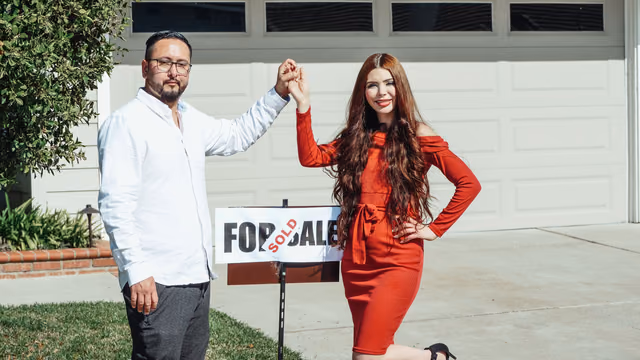 A couple stands hand in hand in front of a house beside a for-sale sign marked sold.