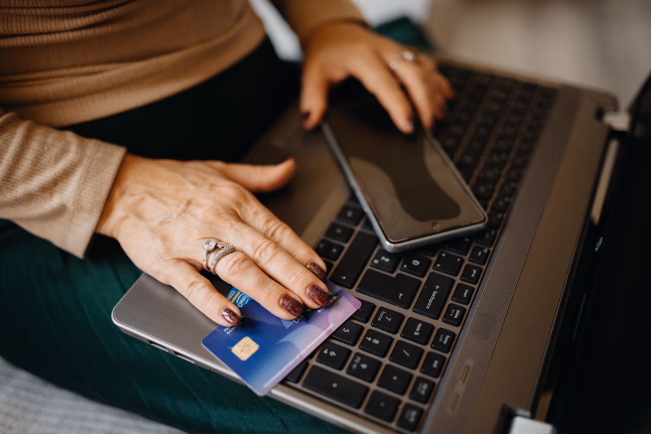 Two hands resting on a laptop keyboard, one holding an unbranded purple credit card, a phone beside them