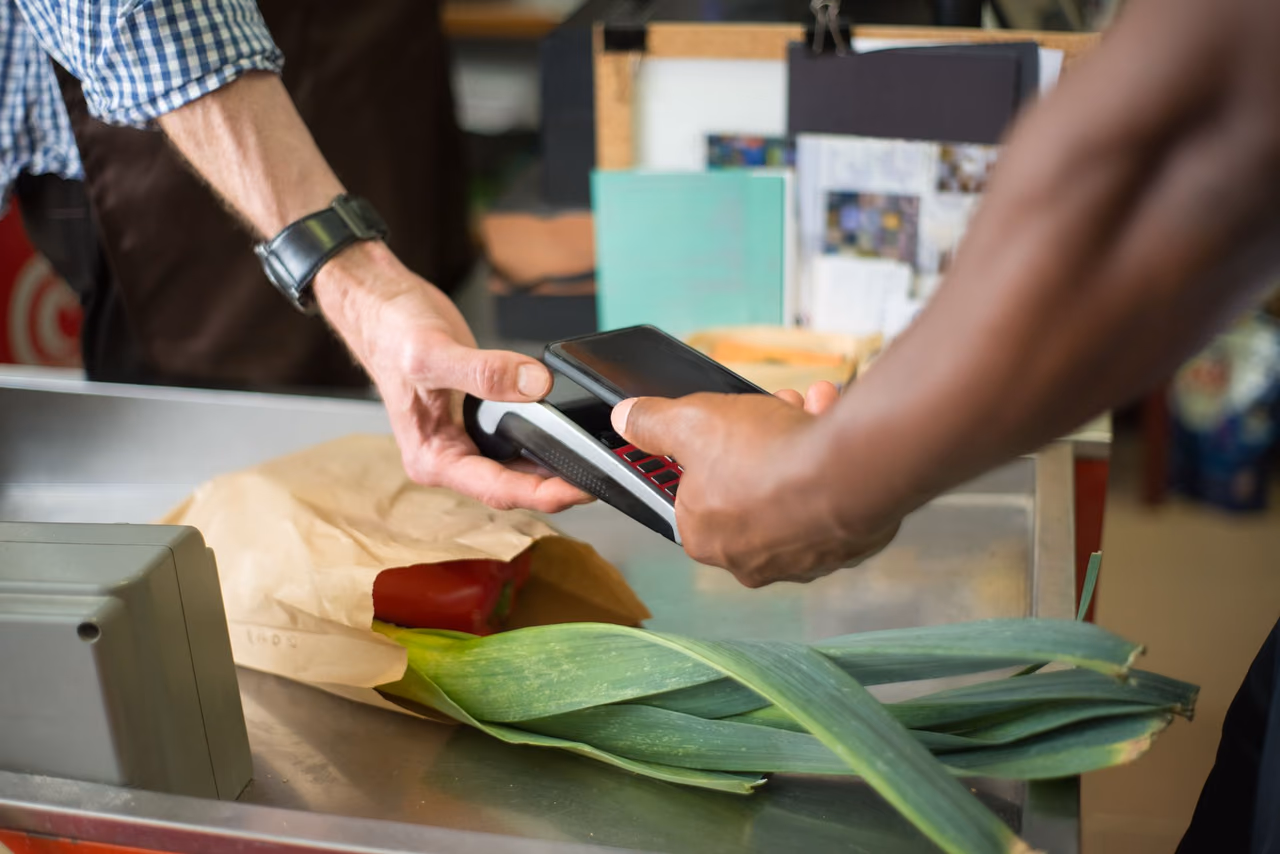 A customer holds a phone over a card reader at a grocery counter while the cashier steadies the reader, with leeks and a paper bag in front