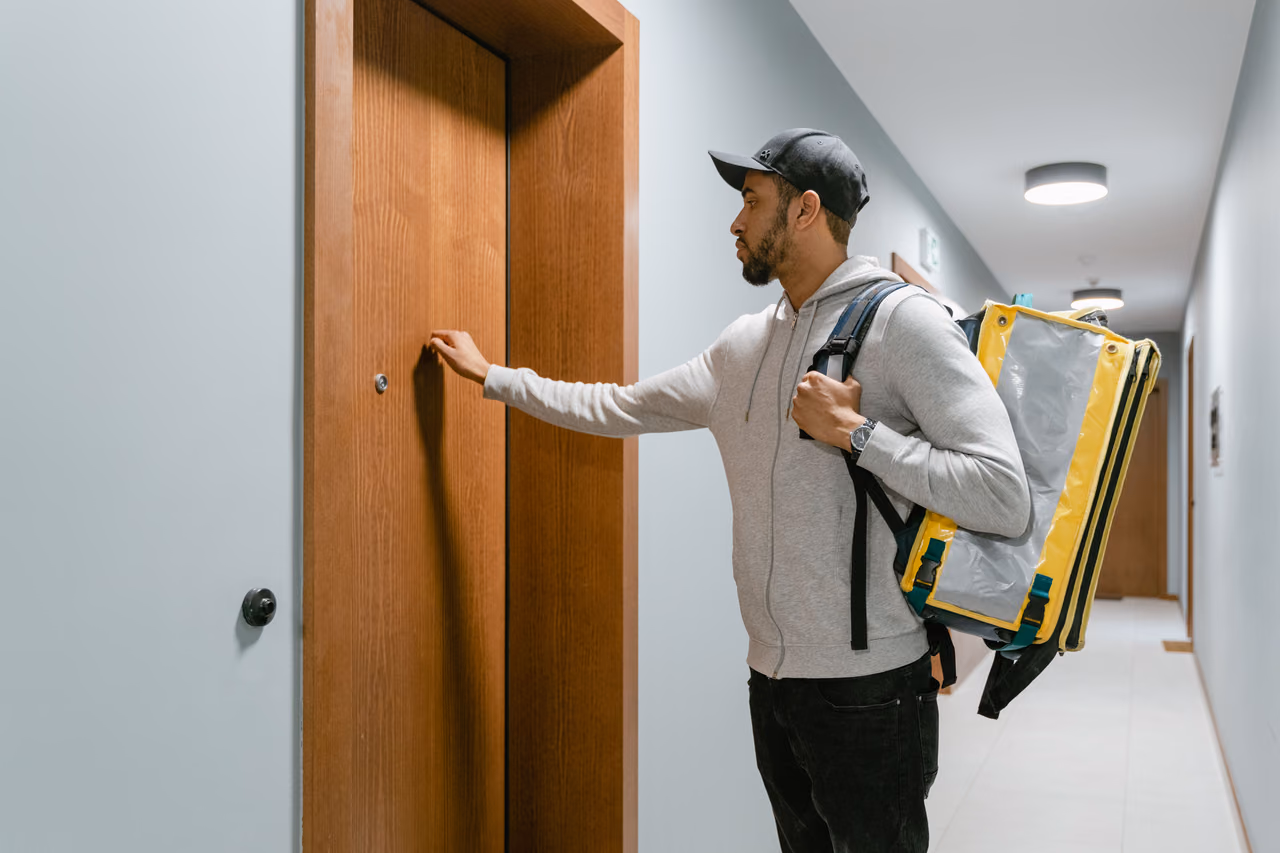 A delivery courier with a yellow insulated backpack knocks on an apartment door in a hallway