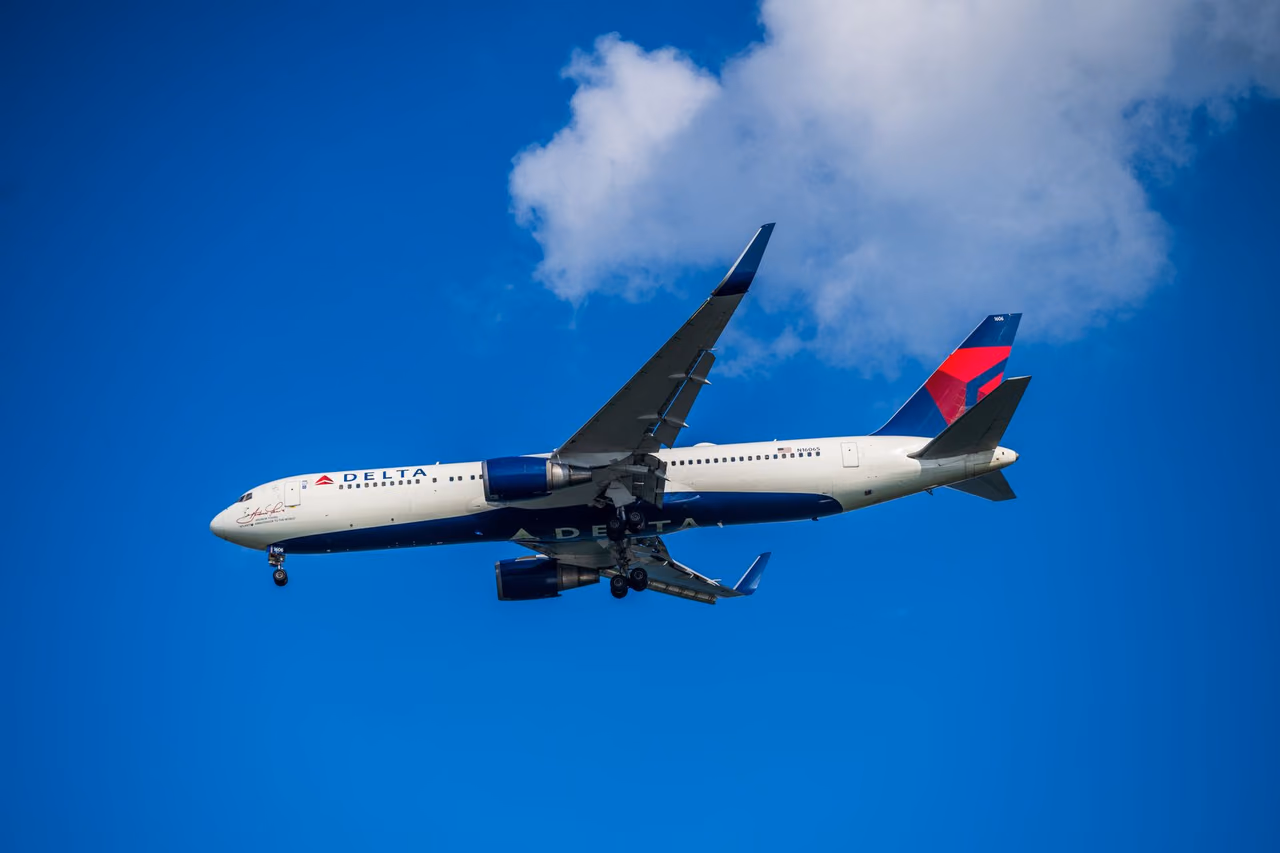 A Delta Air Lines airliner descending with its landing gear down against a deep blue sky and one white cloud