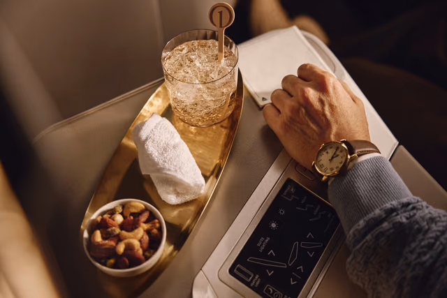 A traveler's hand rests beside the seat controls in a Delta One suite, next to a gold tray holding an iced drink with a Delta One stirrer, a rolled hot towel and a bowl of mixed nuts