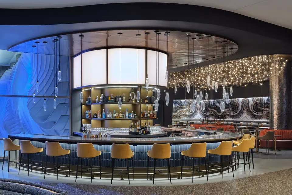 Curved bar with mustard-colored stools, hanging glass pendant lights and a ceiling of string lights inside the Delta Sky Club in Concourse B at Salt Lake City