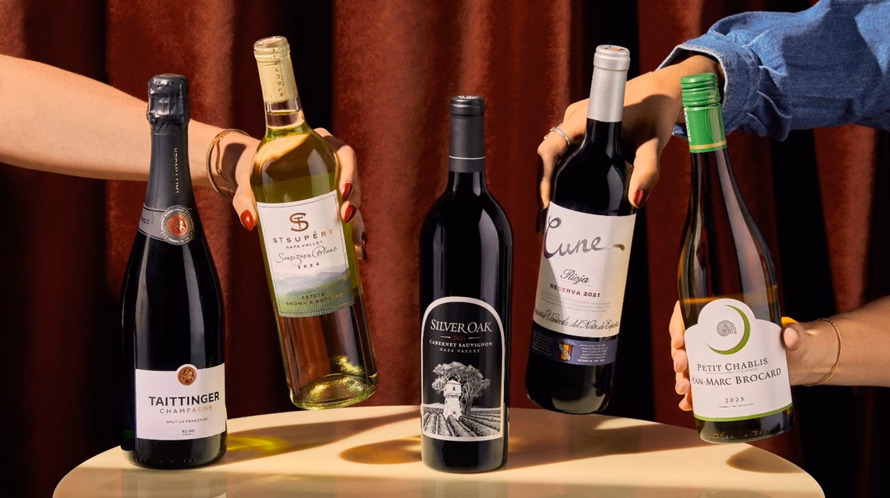 Five wine bottles, including Taittinger Champagne and Silver Oak Cabernet Sauvignon, standing on or held above a small round table