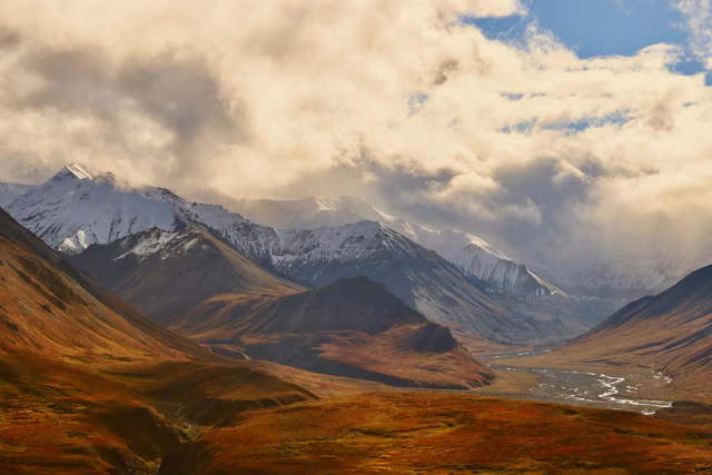 A braided river runs through an autumn-colored valley below snow-capped mountains and heavy clouds in Denali National Park, Alaska