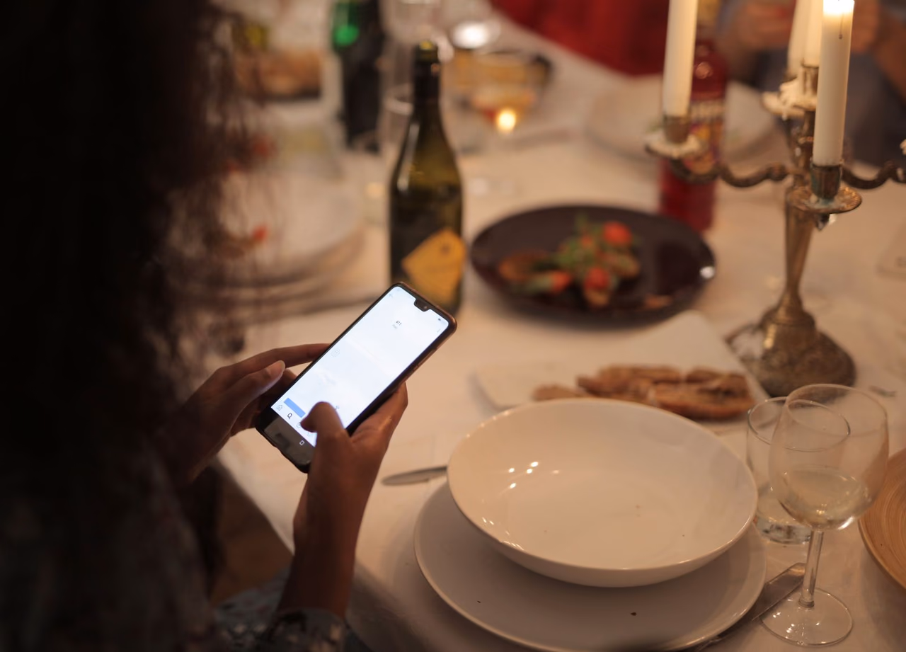 A diner holds a smartphone over a candlelit restaurant table set with plates, a wine bottle and glasses