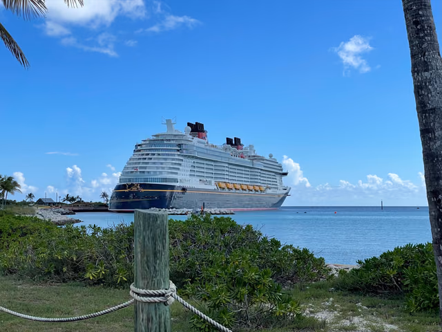 The Disney Wish, a navy-hulled cruise ship with red funnels, docked at Disney Castaway Cay, seen past palm trees and beach shrubs