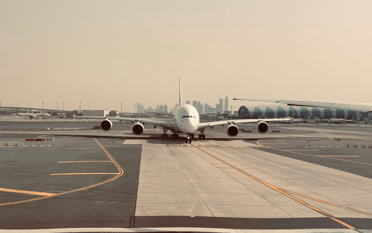 A four-engine double-deck airliner taxiing head-on at Dubai International Airport, with a curved terminal concourse and the hazy Dubai skyline behind it