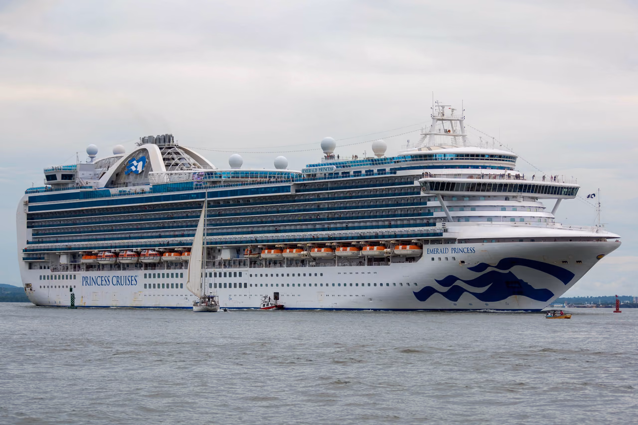 The Emerald Princess cruise ship under way on gray water beneath an overcast sky, with a small sailboat passing alongside