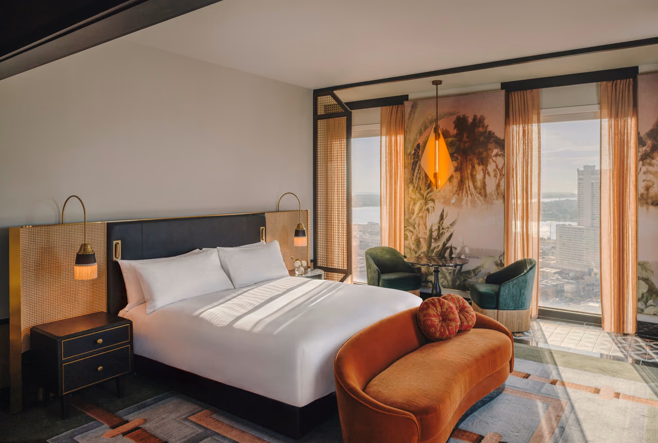 A Deluxe River View guest room at Fairmont New Orleans with a large white bed, an orange velvet settee and floor-to-ceiling windows over the city