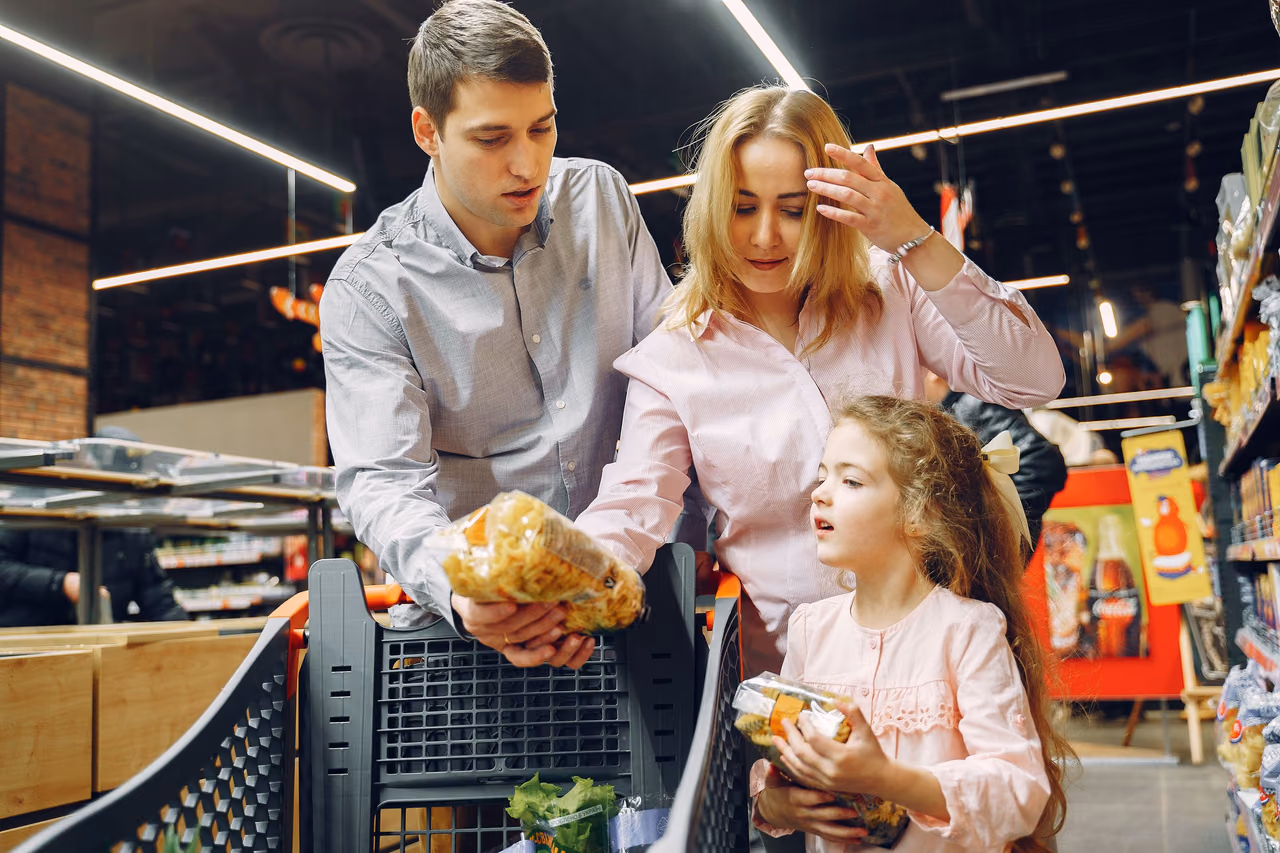 A family with a young daughter choosing groceries beside a shopping cart in a supermarket aisle.