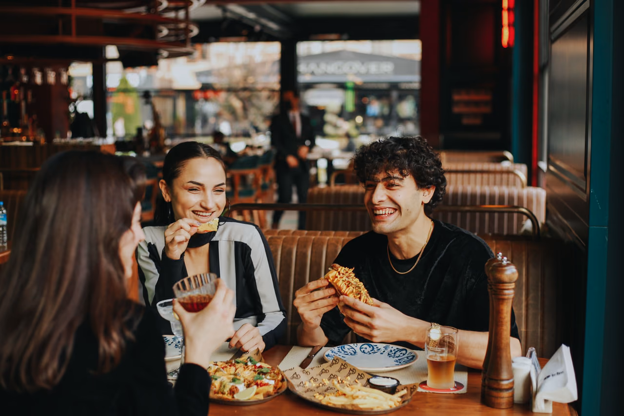 Three friends laughing over plates of food, fries and drinks at a booth table in a busy restaurant