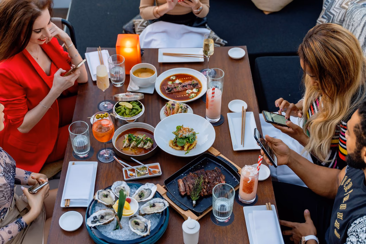 Diners holding phones around a restaurant table set with oysters, a steak and several plates, seen from above