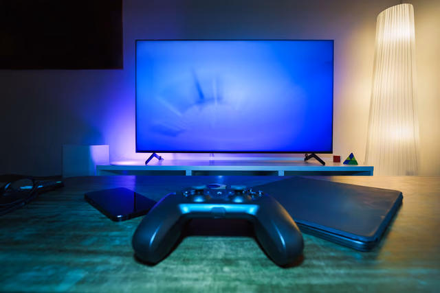 A game controller on a wooden desk in front of a glowing blue TV, with a tablet and a closed laptop beside it in a dim living room