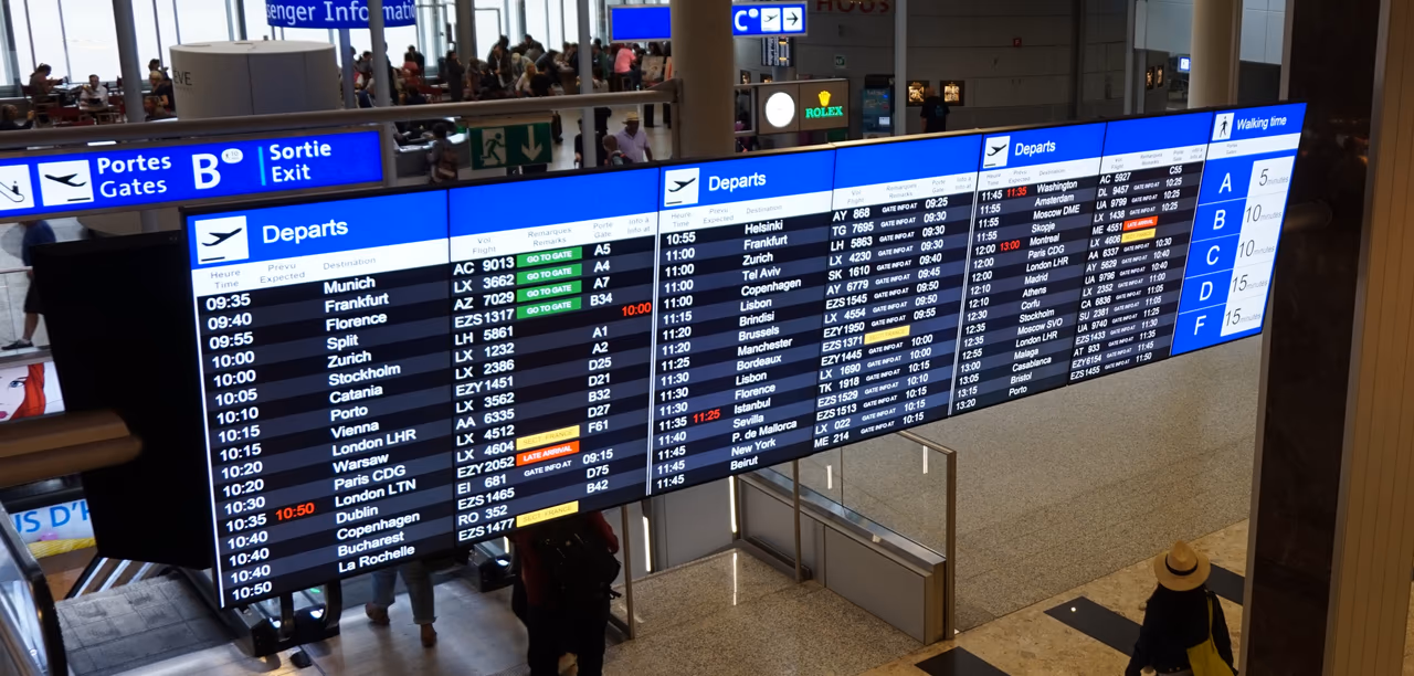 A row of blue departure boards above a terminal walkway at Geneva Airport, listing flights to European cities