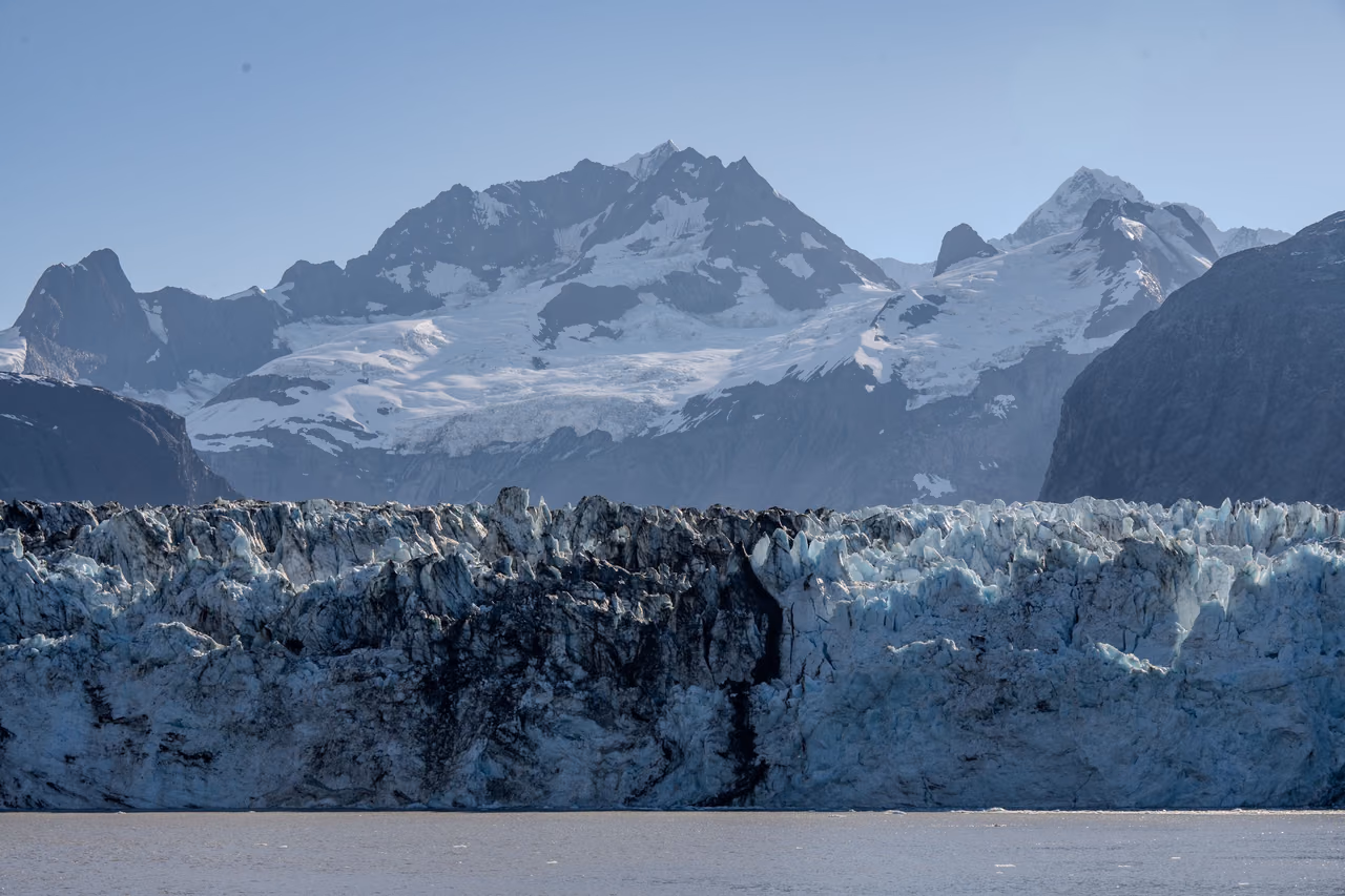 The jagged blue and gray ice face of a tidewater glacier at the waterline, with snow-covered peaks behind it under a clear sky