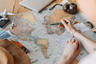 Hands marking a route with a pen on a vintage world map, with a toy airplane, a passport, a camera and a laptop on a wooden table