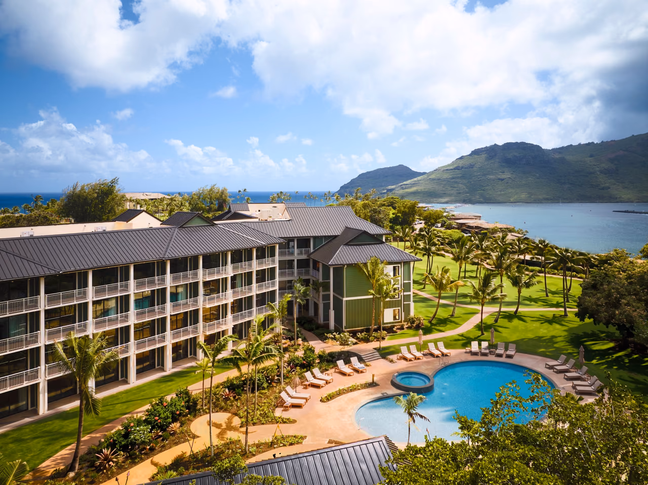 Aerial view of Hale Hokuala Kauai's pool and lounge chairs beside its low-rise guest wings, with palm-lined lawns, Nawiliwili Bay and green mountains behind