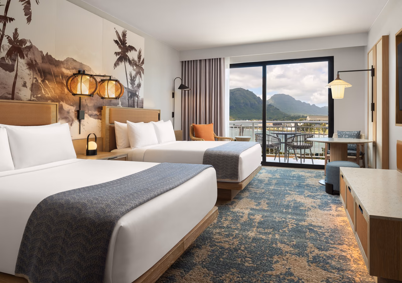 Guest room with two queen beds, a palm tree and mountain mural behind the headboards, and a sliding glass door to a balcony facing green mountains and the bay