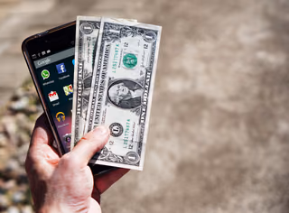 A hand holding two U.S. one-dollar bills in front of a smartphone home screen