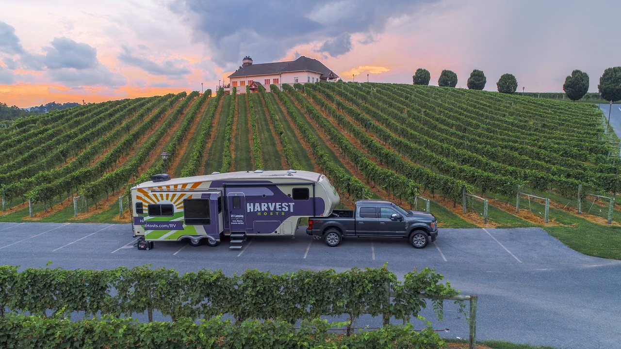 A Harvest Hosts fifth-wheel trailer hitched to a gray pickup truck, parked beside rows of grapevines below a large hilltop building at sunset