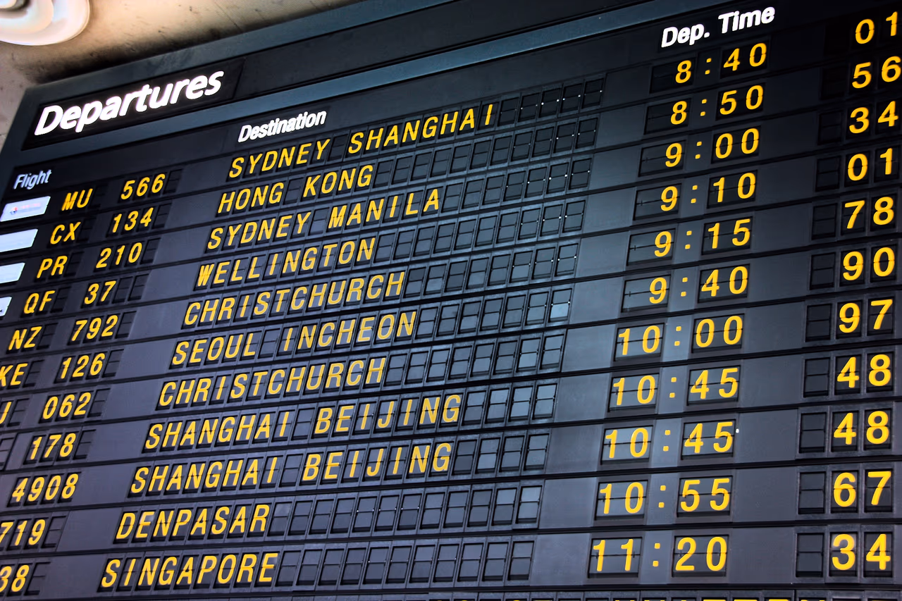 A split-flap airport departures board in yellow lettering on black, listing flights to Sydney, Shanghai, Hong Kong, Manila, Wellington, Christchurch, Seoul Incheon, Beijing, Denpasar and Singapore with their departure times