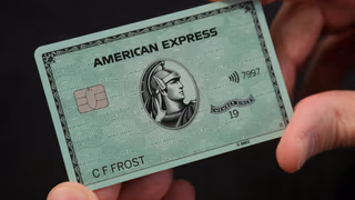 A hand holding a pale green American Express card, its centurion portrait, chip and contactless symbol visible, against a dark background
